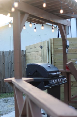 Una pérgola de madera con luces colgantes, una parrilla y una mesa de madera.