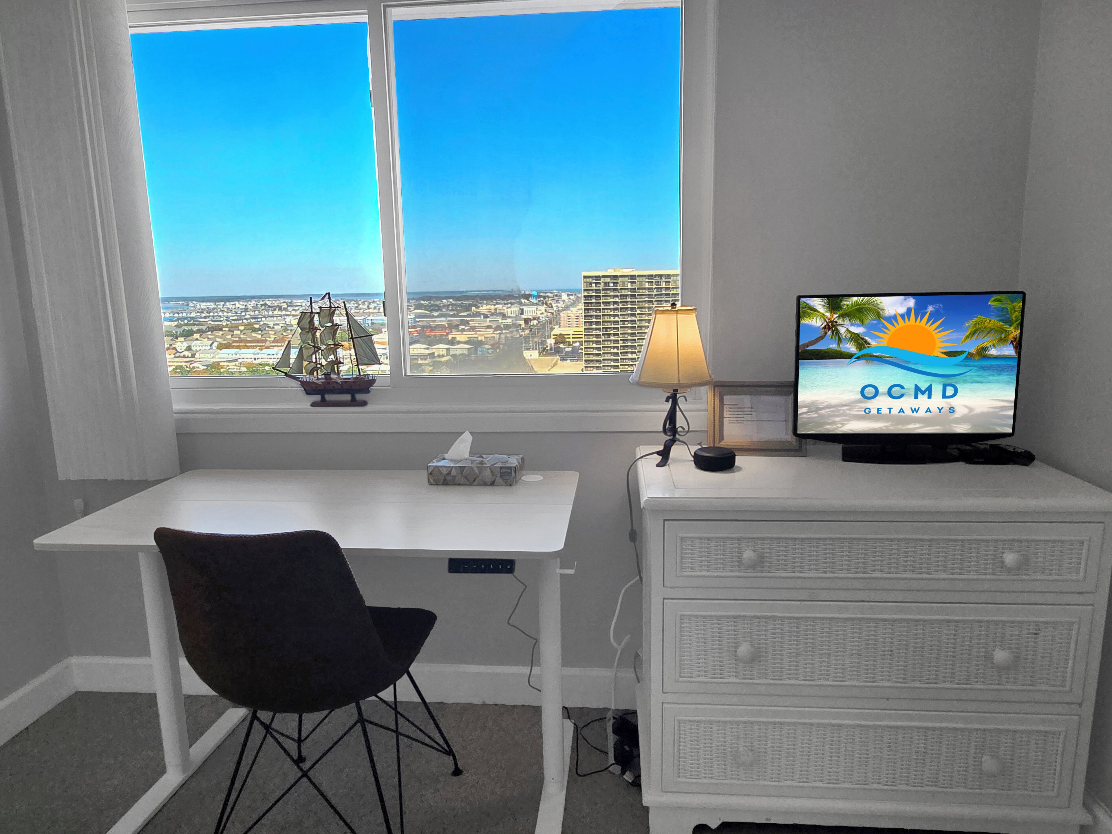 Habitación con escritorio, silla y televisor mostrando 'OCMD GETAWAYS'. Una ventana ofrece vista a la ciudad.