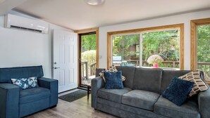 A living room with a grey sofa, a blue armchair, and a white door. There are large windows with a view of greenery outside.