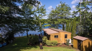 A small wooden cabin with a dock, surrounded by lush greenery and a lake.