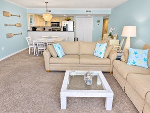 A living room with a beige sofa, a coffee table, and a dining area with white chairs.