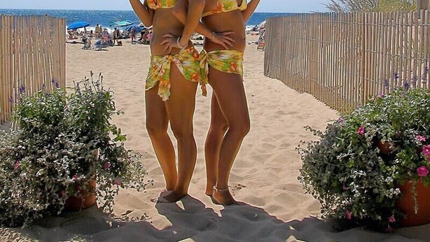 Two women in bikinis carrying trays with drinks and a bottle on a sandy beach with a wooden fence and potted plants. - Ocean Block, 2 Bedroom Condo on 37th St. Sovereign Sea. Next to Coconuts Beach Bar and many other great places! Sovereign Sea #302 (Ocean City)