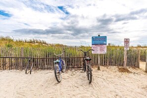 A sandy beach with a wooden fence and two bicycles parked near it. There are signs indicating dune crossing and no parking at the head of the street.