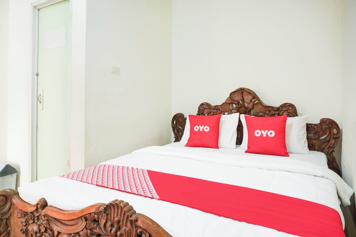 Una cama con cabecera de madera, dos almohadas rojas con la palabra "OYO" y una colcha blanca con manta de estampado rojo y blanco.