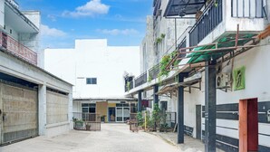 Une rue étroite en milieu urbain, bordée de bâtiments des deux côtés, avec des balcons ornés de plantes et un ciel d’un bleu éclatant.