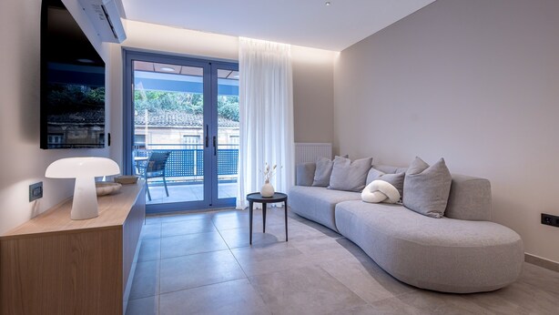 A modern living room with a curved grey sofa, a small round table with a vase, and a mounted TV on the wall. - Phaedrus Living Athens Residences Golden (Athens)