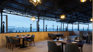 A restaurant with tables set for dining, large windows offering a city view, and hanging light fixtures. - Bogishamol Hotel (Samarkand)