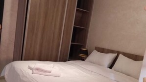 Cama bem arrumada com lençóis brancos e toalha dobrada, guarda-roupa de madeira e mesa de cabeceira com abajur.