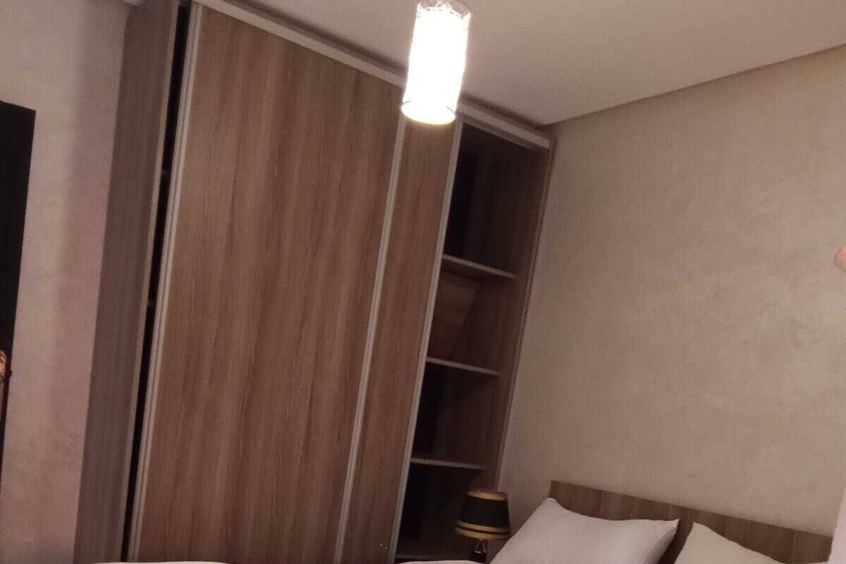 A neatly made bed with white linens and a folded towel, a wooden wardrobe, and a bedside table with a lamp.