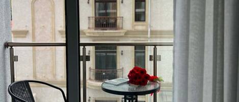 Un balcón con una mesa y sillas, un arreglo floral rojo y un edificio con tejado de tejas rojas al fondo.