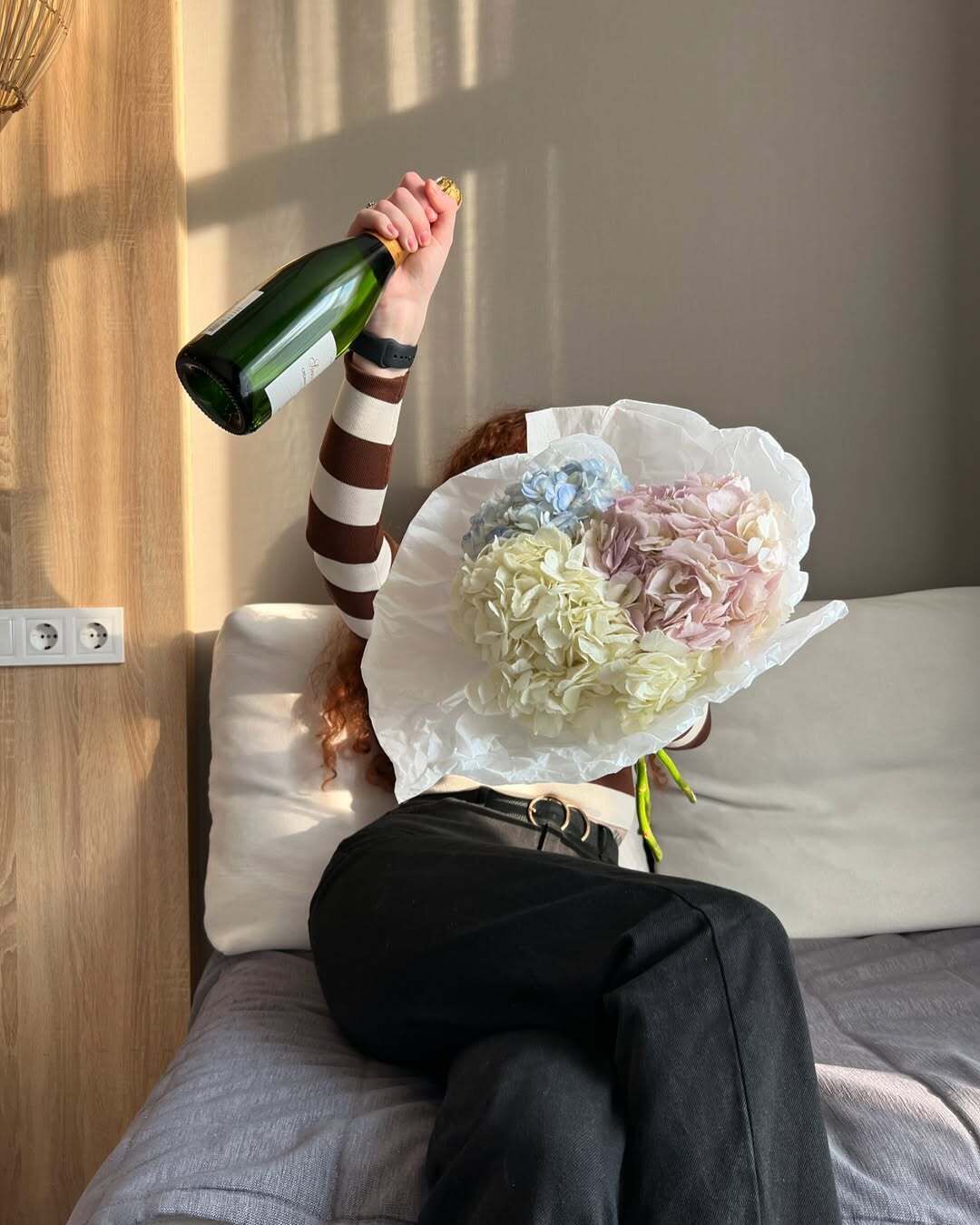 A person sitting on a bed with a bouquet of flowers and a bottle of champagne.