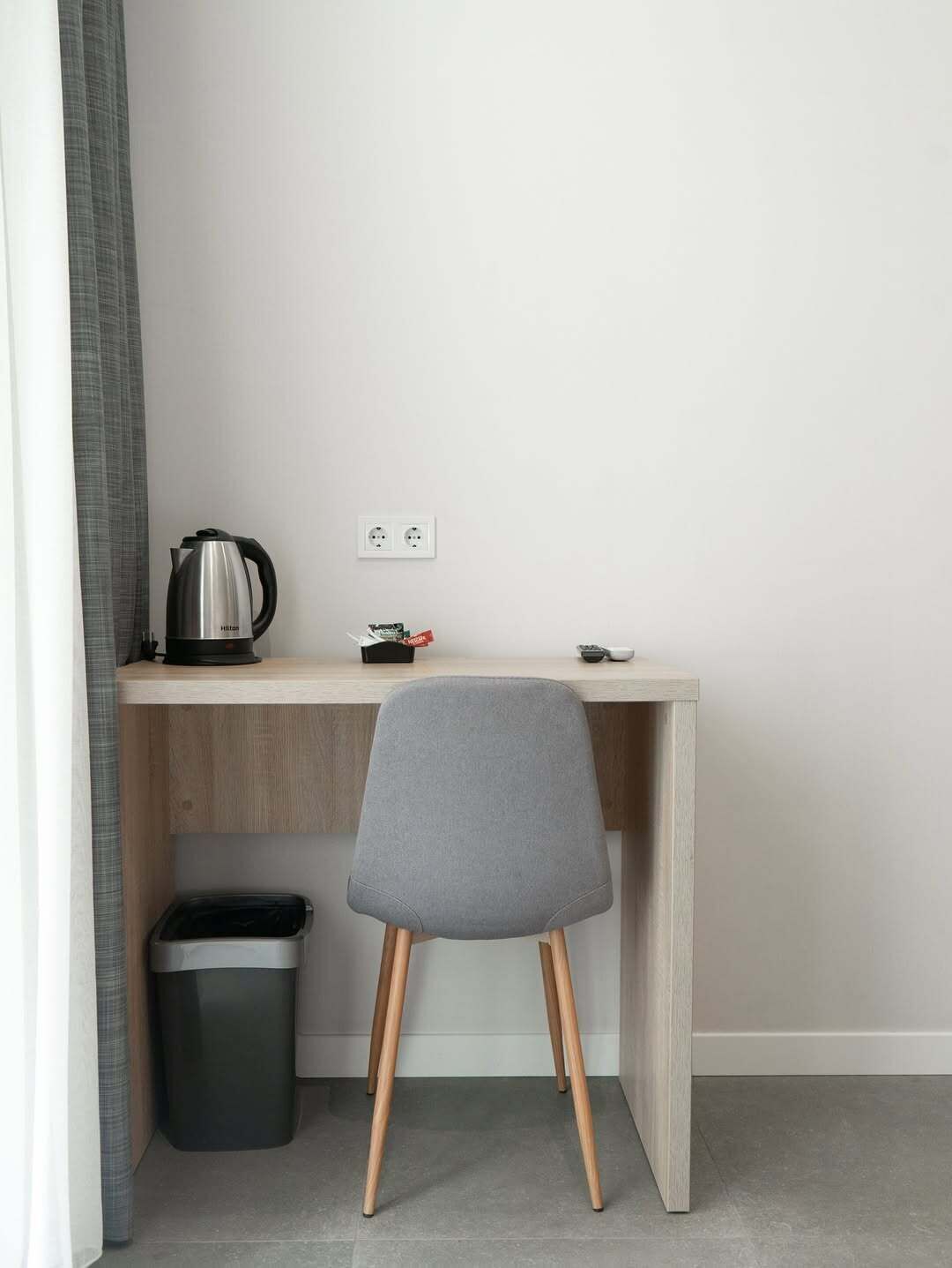 Un espacio de trabajo pequeño y moderno con un escritorio de madera, una silla gris, una tetera y un cubo de basura.