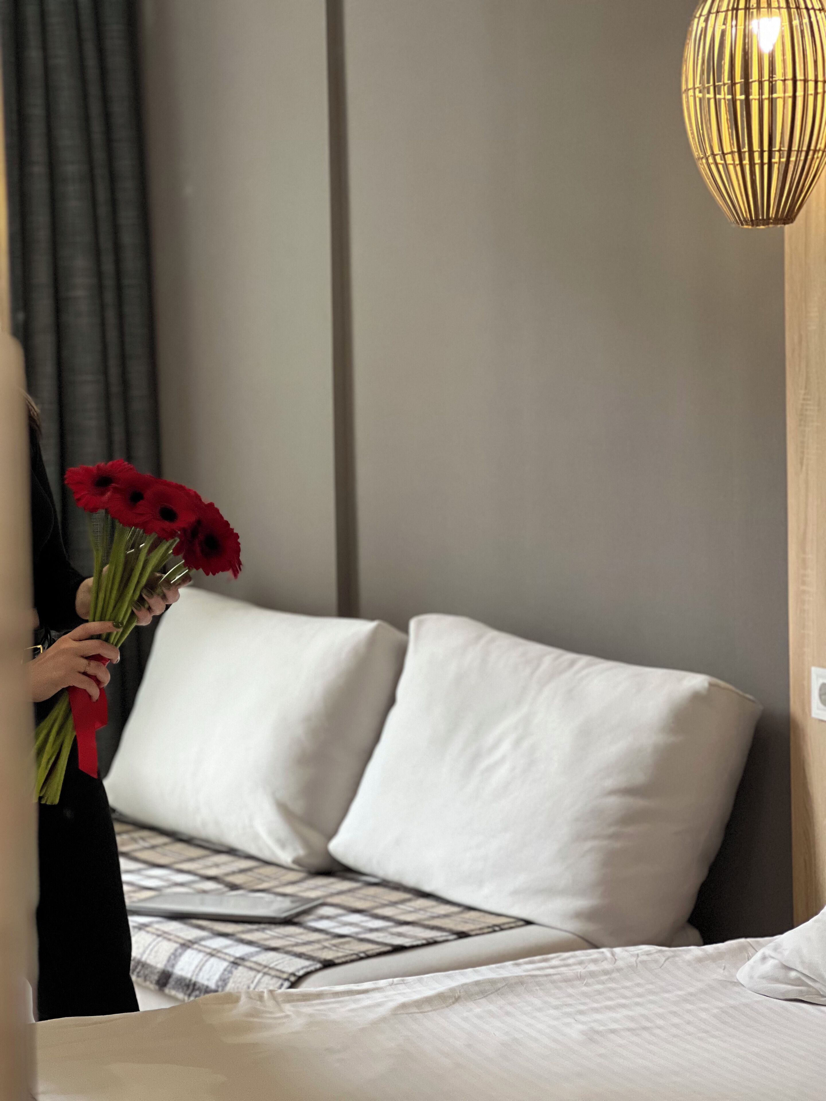 Habitación de hotel con cama, almohadas, lámpara de noche y una persona sosteniendo un ramo de flores rojas.