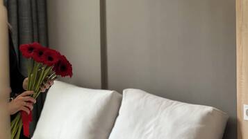 Habitación de hotel con cama, almohadas, lámpara de noche y una persona sosteniendo un ramo de flores rojas.