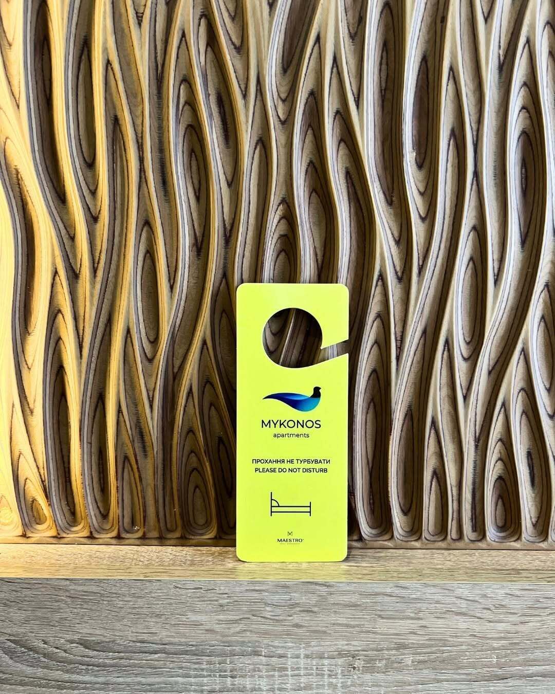 A yellow sign with the text 'MYKONOS apartments' and a bird logo, placed against a wooden wall with a wavy pattern.