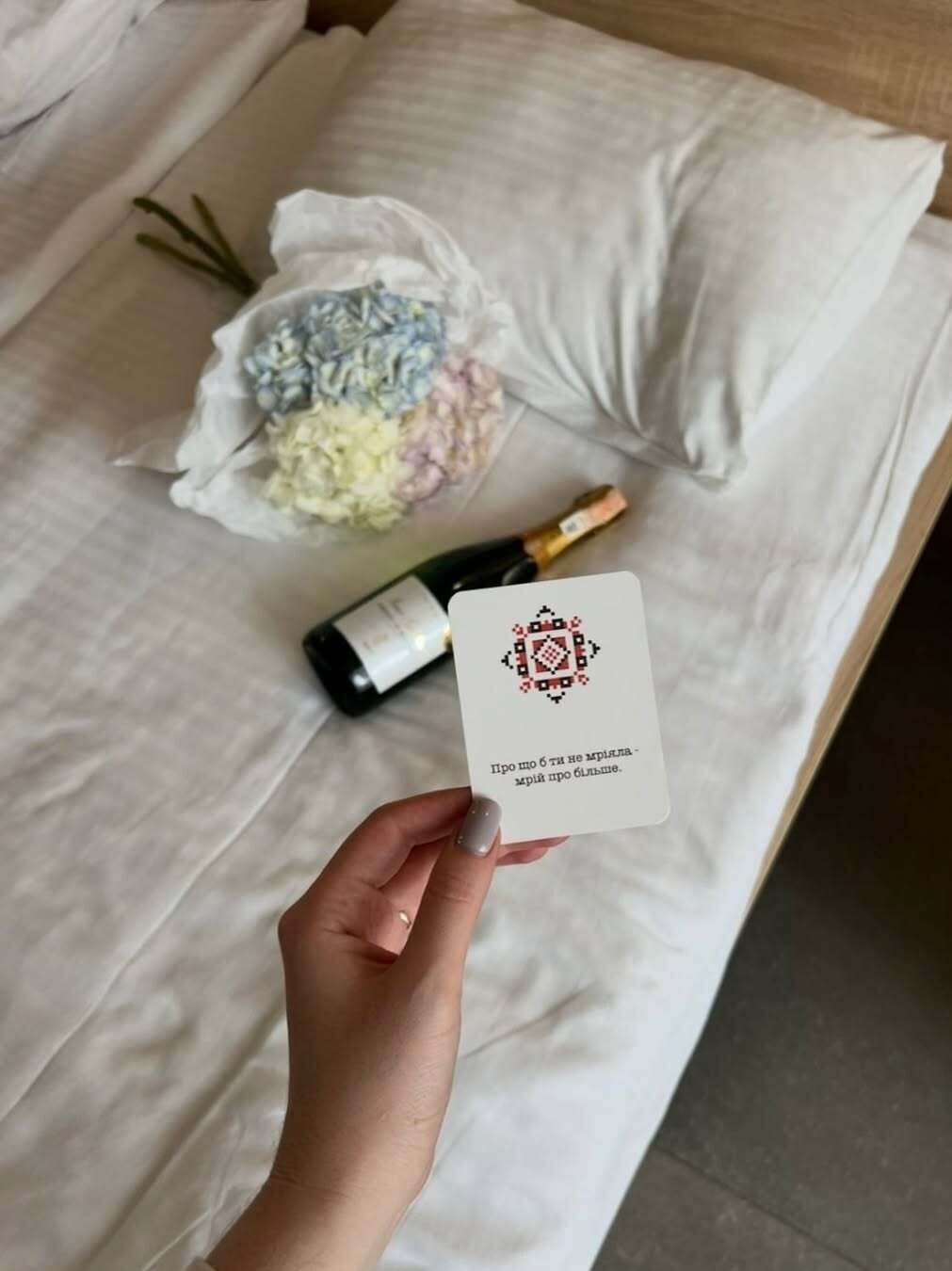 Eine Hand hält eine Karte mit kyrillischer Schrift, eine Flasche und einen Blumenstrauß auf einem Bett.