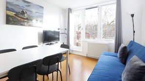 Un salon moderne avec un canapé bleu, une table à manger et des chaises, un téléviseur à écran plat et une grande fenêtre donnant sur des arbres et des immeubles.