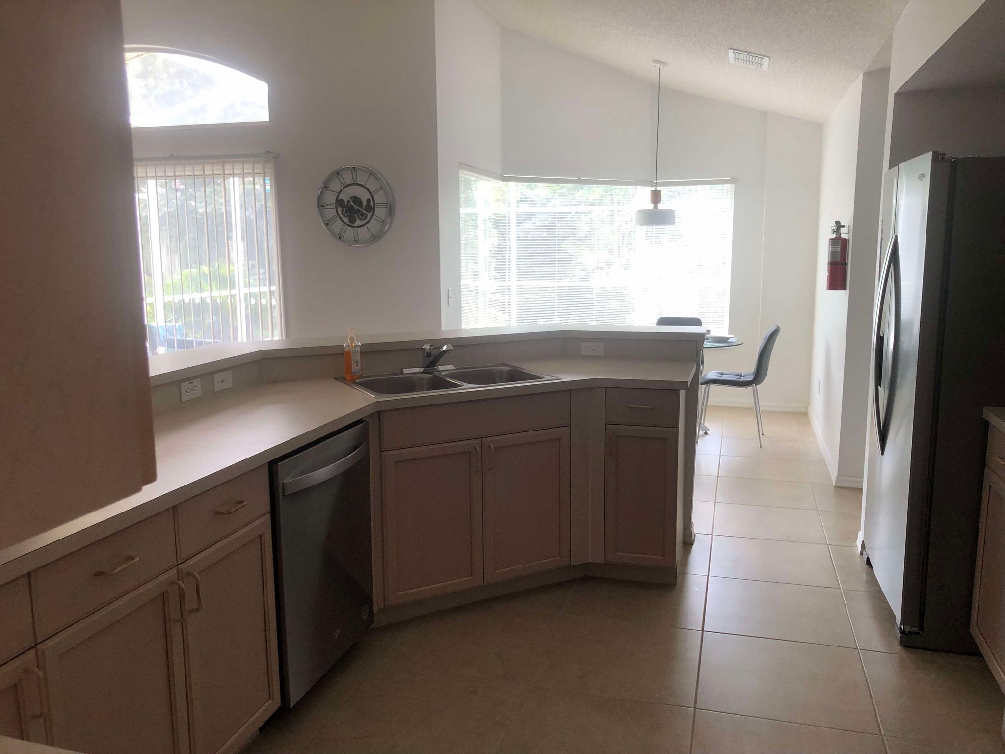 A kitchen with a dishwasher, a sink, and a refrigerator. There is a window with blinds, a clock on the wall, and a chair in the corner.