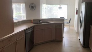 Eine Küche mit Geschirrspüler, Spüle und Kühlschrank. Ein Fenster mit Jalousien, eine Uhr an der Wand und ein Stuhl in der Ecke.
