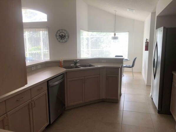 A kitchen with a dishwasher, a sink, and a refrigerator. There is a window with blinds, a clock on the wall, and a chair in the corner.