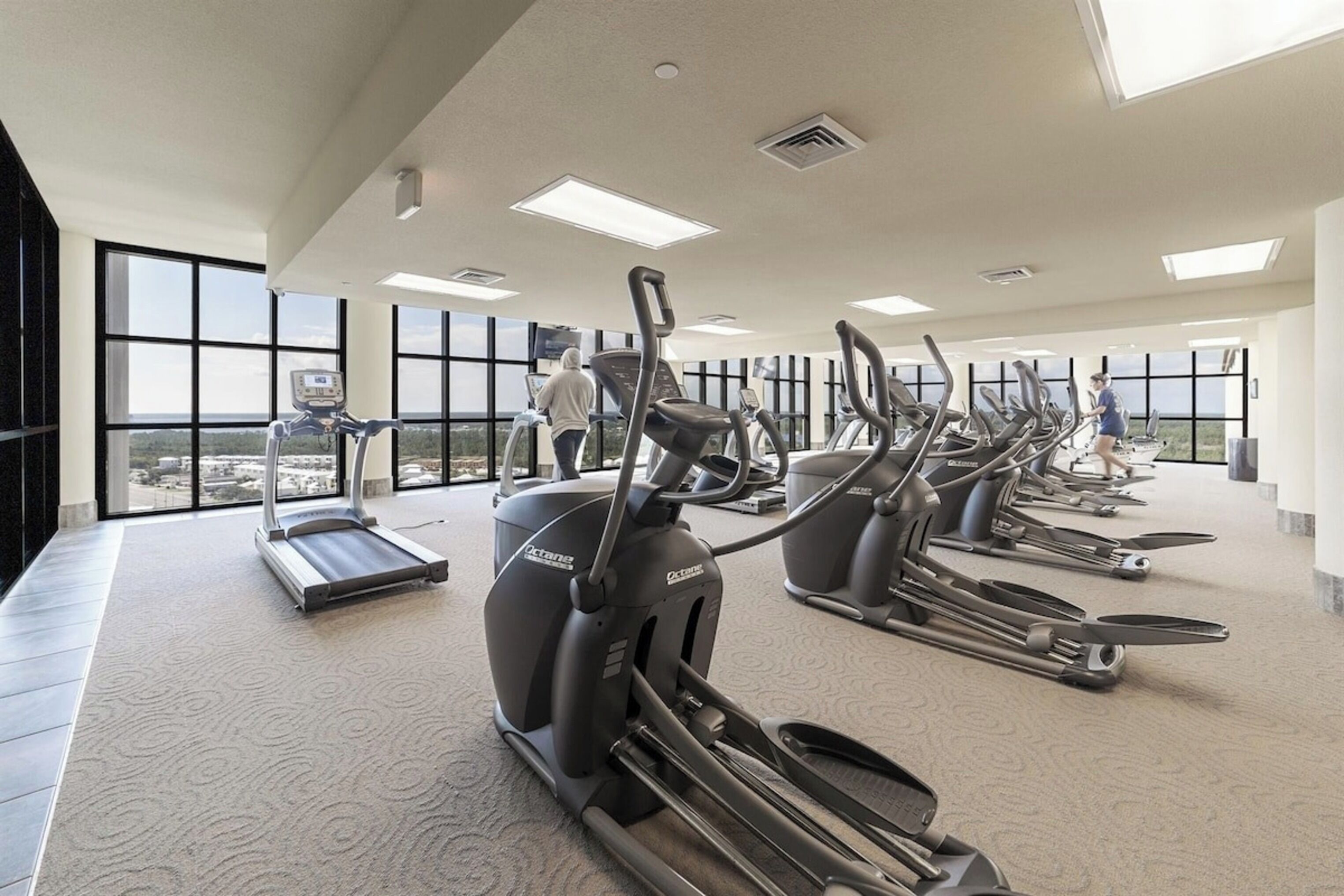 A gym with treadmills and elliptical machines, large windows, and a high ceiling.