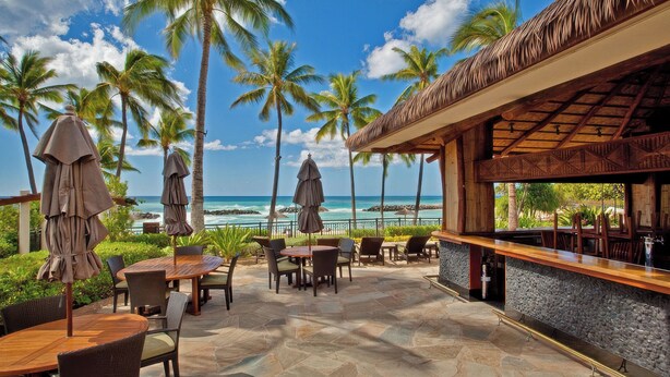 An outdoor patio with tables and chairs, a bar area, and a view of the ocean and palm trees.