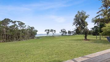 Un espacio al aire libre con césped, una terraza de madera, árboles y vista al agua.