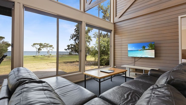 A modern living room with a large window overlooking a beach, a leather sofa, a small table, and a TV on the wall. - Avon - Waterfront Retreat: Secluded lodges perfect for nature lovers (Forge Creek)