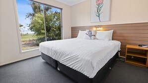A modern bedroom with a large bed, a wooden headboard, a nightstand, and a view of trees and water through a large window. - Avon - Waterfront Retreat: Secluded lodges perfect for nature lovers (Forge Creek)