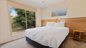 A bedroom with a large bed, a window with a view of trees, a wooden headboard, and a small table.