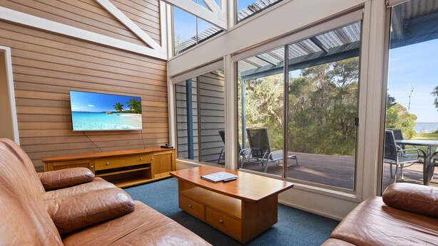 A spacious living area with leather sofas, a wooden coffee table, and a TV mounted on the wall. Large windows offer a view of the outdoors. - Dargo - Waterfront Retreat: Secluded lodges perfect for nature lovers (Forge Creek)