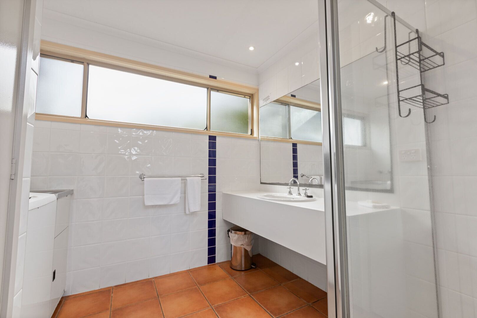 Une salle de bain moderne avec une douche à parois de verre, des murs recouverts de carreaux blancs, une bande de carreaux bleus en guise d’élément décoratif, une fenêtre au-dessus du lavabo et une poubelle visible.