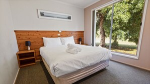 Una cama bien hecha con sábanas blancas, un cabecero de madera y una mesita de noche con una lámpara. Un ventanal ofrece vistas a árboles y un césped.