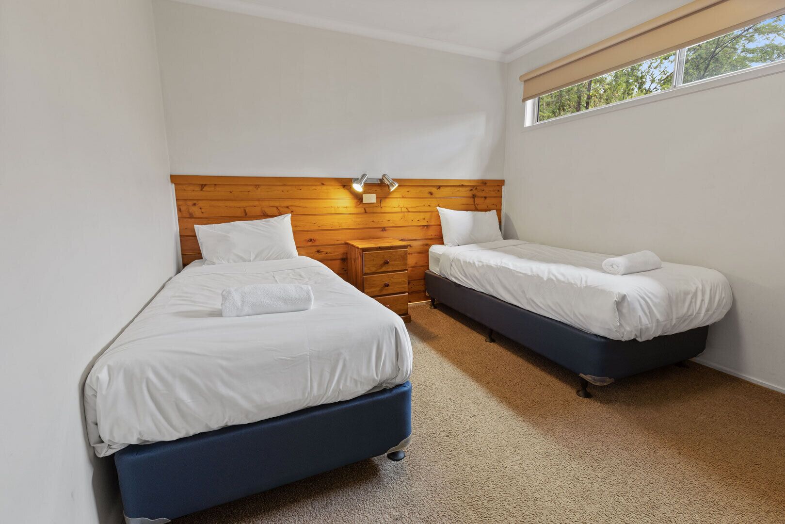 Quarto com duas camas, uma cabeceira de madeira, mesa de cabeceira e janela com vista para árvores.