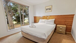 A bedroom with a large bed, a wooden headboard, a nightstand, and a window with a view of trees. - Westwind - Waterfront Retreat: Secluded lodges perfect for nature lovers (Forge Creek)