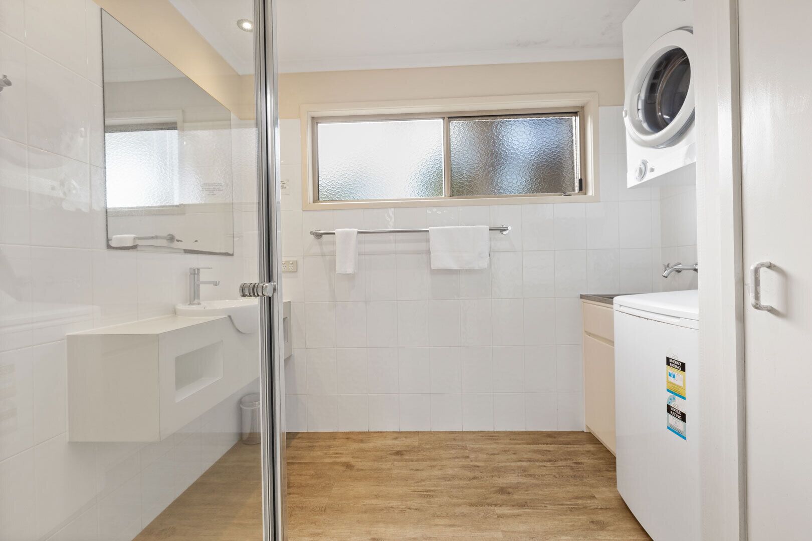A modern bathroom with a glass shower enclosure, a built-in sink, a window above the shower, and a washing machine.