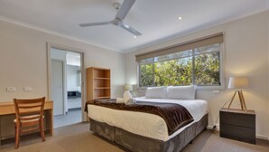 Une chambre à coucher comprenant un lit, un bureau, une chaise, une bibliothèque, une fenêtre donnant sur des arbres et un ventilateur de plafond.