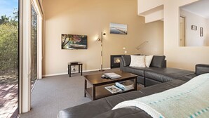 Een moderne woonkamer met een bank, salontafel en open haard. Er hangt een schilderij aan de muur en een raam met uitzicht op bomen.