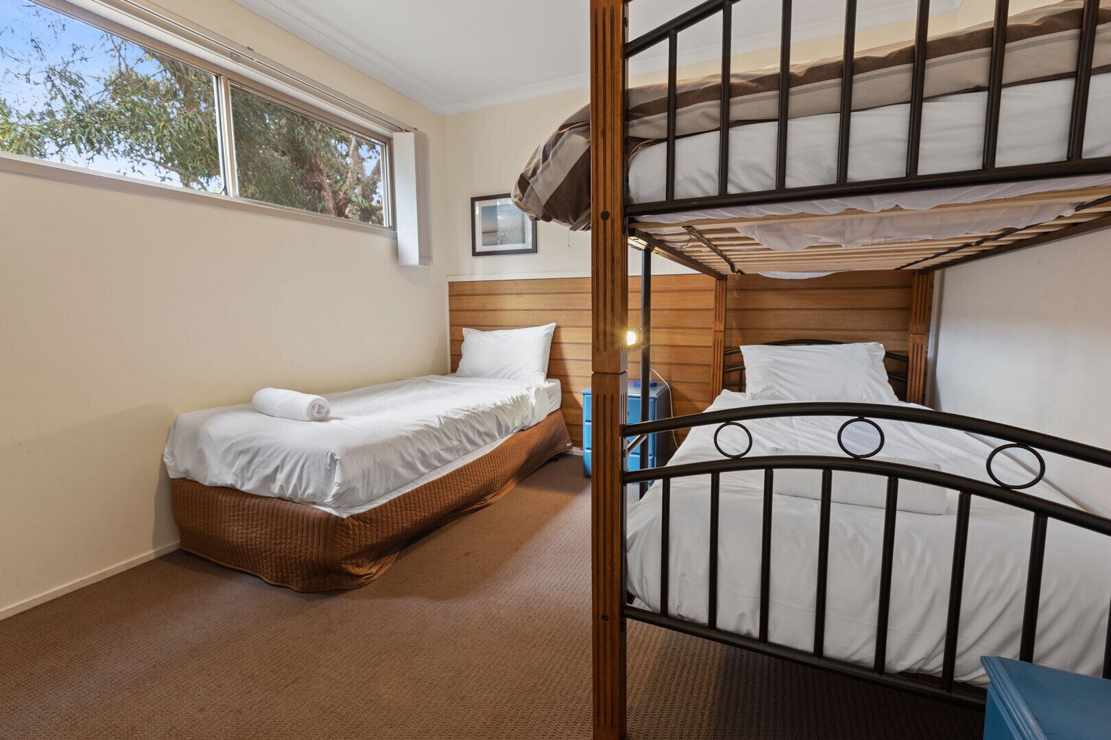 A room with two beds, one with a wooden headboard and the other with a metal frame. There is a window providing natural light and a small table with a blue chair.