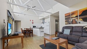 A modern living room with a sofa, coffee table, and TV. There is a dining area with chairs and a kitchen in the background. - Secluded lodges perfect for nature lovers (Forge Creek)