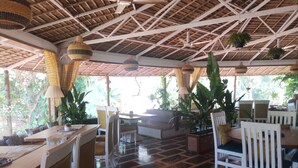 Un comedor interior con mesas y sillas de madera, lámparas colgantes y plantas en macetas.