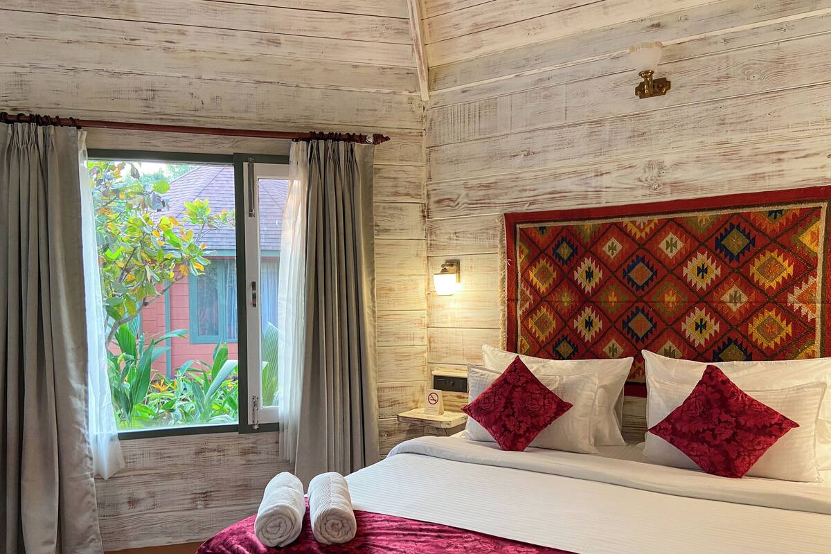 A hotel room with a wooden ceiling, a large bed with white linens, red and white pillows, and a view of greenery outside.