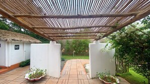 Un pasillo cubierto con techo de paja, plantas en macetas y un edificio blanco con un letrero que indica el baño de hombres.