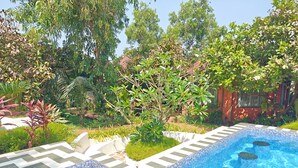Una piscina con borde de azulejos, rodeada de exuberante vegetación y un edificio rojo al fondo.