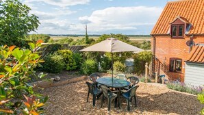 A patio with a table and chairs, surrounded by a garden and a brick house with a red roof. - Host & Stay | Laylands Yard (Wells-next-the-Sea)