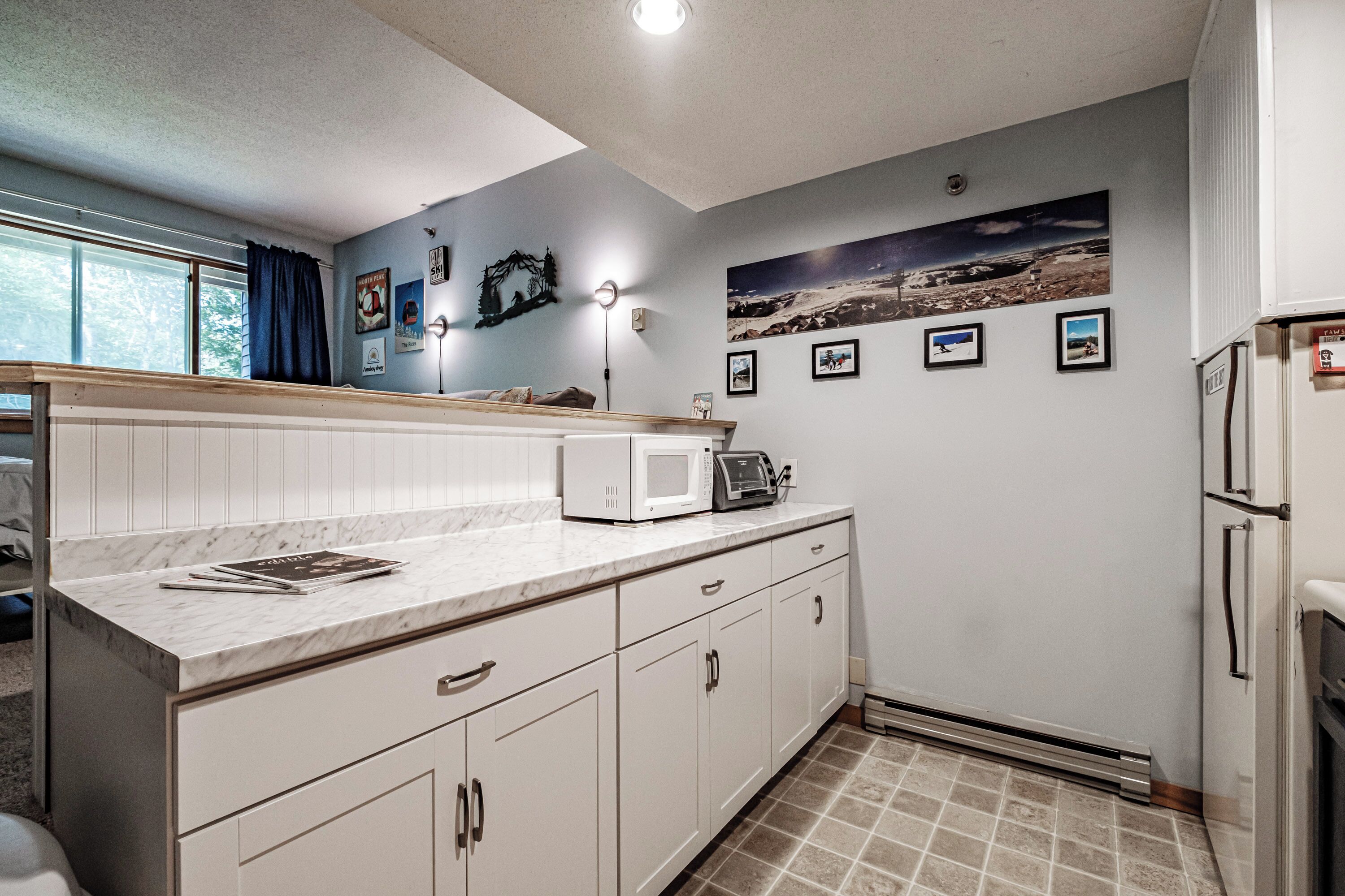 A kitchen with white cabinets, a marble countertop, a microwave, and a refrigerator. There are framed pictures on the wall and a window with curtains.