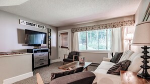 A living room with a television, a wooden coffee table, a sofa, and a chair. There is a window with curtains and a sign that reads "SUNDAY RIVER".