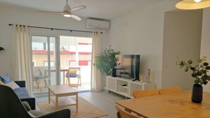 Ett vardagsrum med en soffa, ett matbord och en tv som står på ett vitt skåp. Det finns en takfläkt, en växt och ett fönster med gardiner.