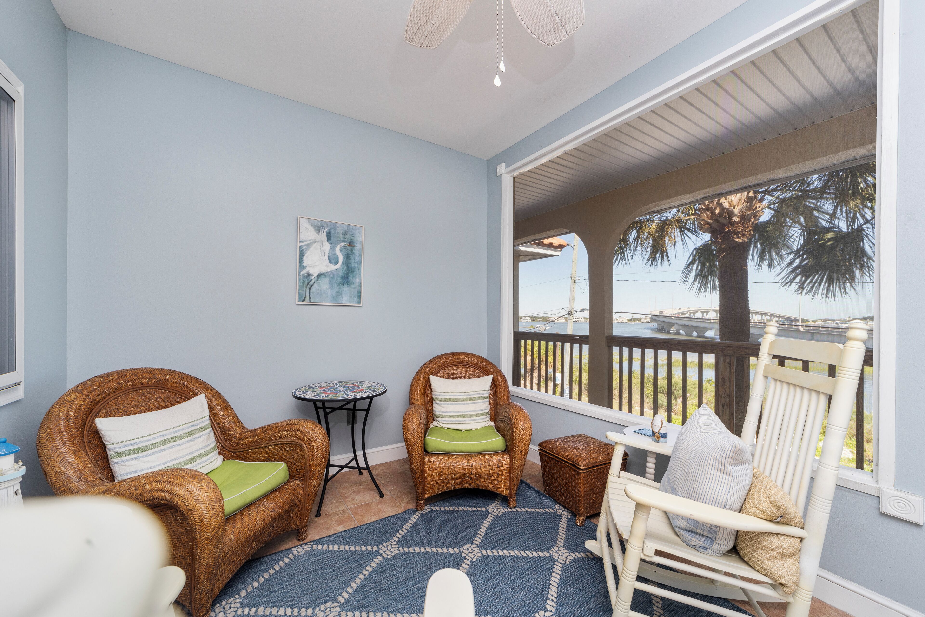 A room with two wicker chairs, one with a green cushion and another with a striped cushion, a small round table, and a view of a bridge and palm trees through a balcony door.
