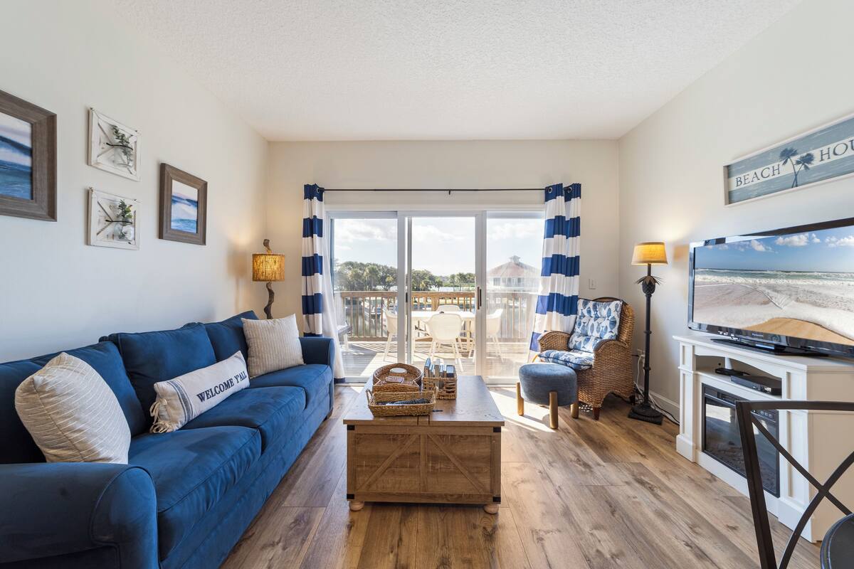 Una sala de estar con un sofá azul, una mesa de centro de madera y un televisor sobre un mueble blanco. Hay un balcón con vista al océano y un letrero que dice "BEACH HOUSE".